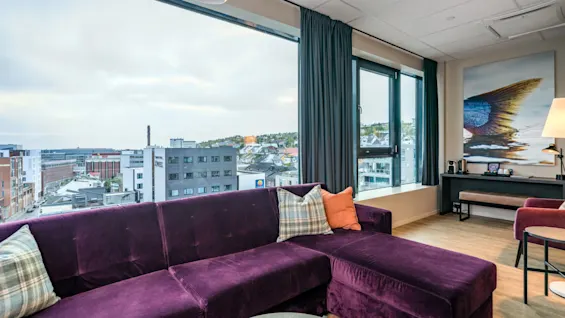 Et moderne oppholdsrom med en stor lilla seksjonssofa og et bredt vindu med utsikt over en by. Kunstverk på veggen.