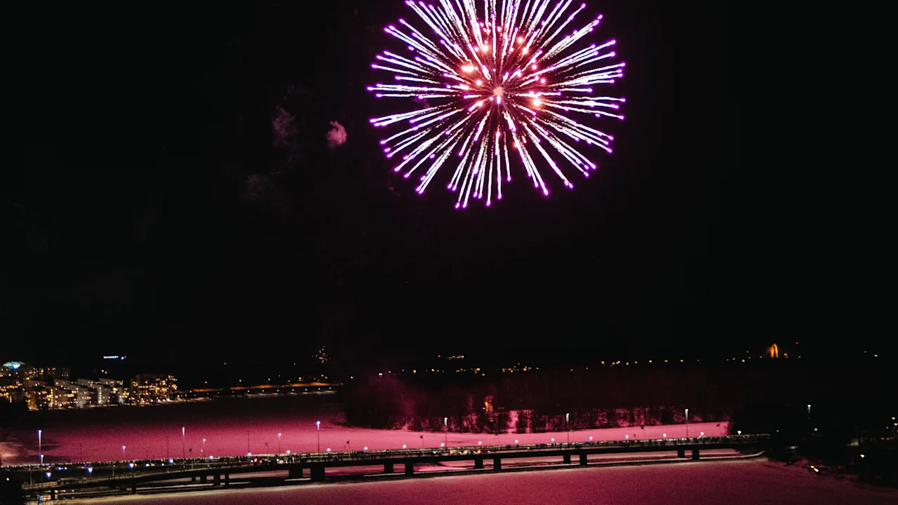 Ett färgstarkt rosa fyrverkeri som exploderar på natthimlen över en bro och vatten som reflekterar det rosa skenet.