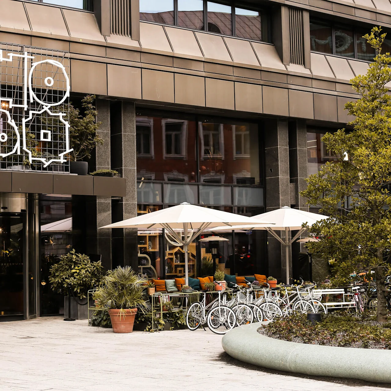 Hotel facade displays geometric Hobo signage above entrance, with potted plants and outdoor seating under umbrellas on a city street.