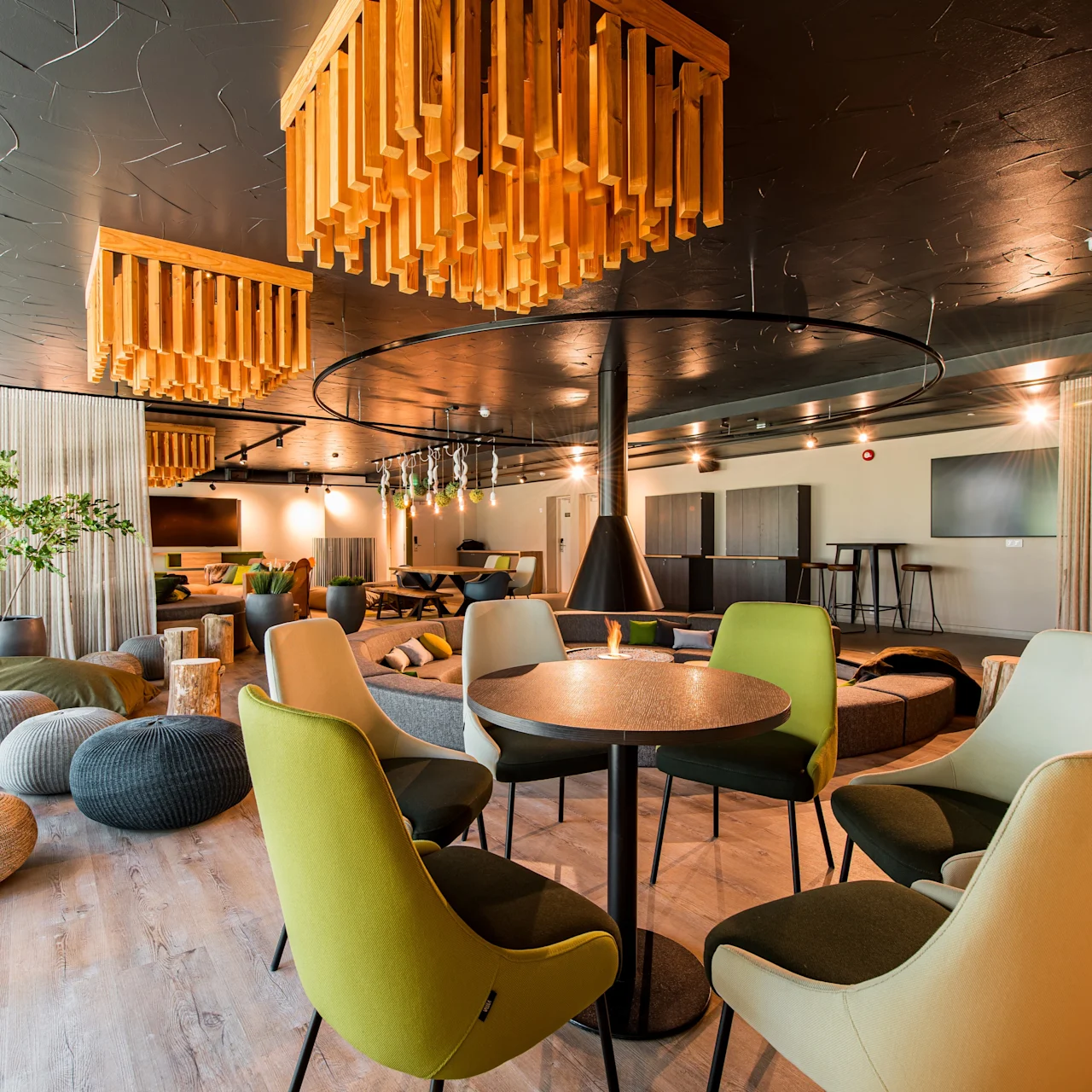 Chairs surround a round table in a modern lounge, featuring wooden ceiling lights and large windows. Soft seating and plants create a cozy atmosphere.