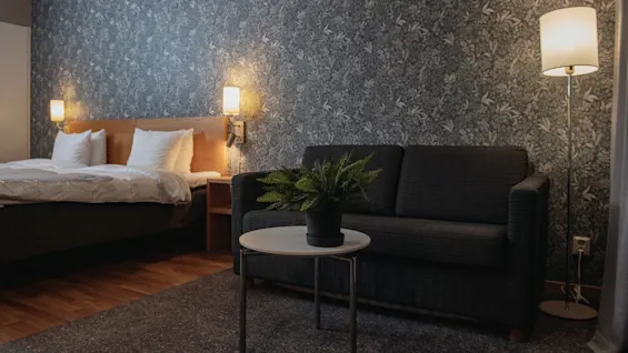 Bed with white pillows adjacent to a dark sofa under a floor lamp; small table with a potted plant sits in a room with floral-patterned wallpaper.