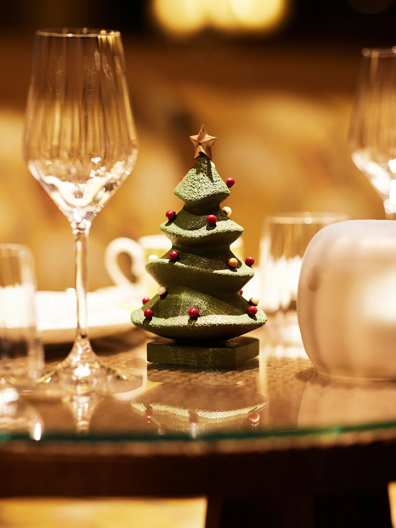 A small decorated Christmas tree ornament sits on a glass table, surrounded by elegant wine glasses in a softly lit setting.