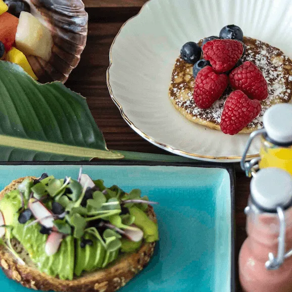 A round pancake topped with raspberries and blueberries sits on a white plate beside a cup of coffee. Nearby, avocado toast and sliced grapefruit complement the breakfast table.
