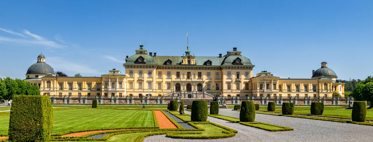 Et storslået gult palads med grønne tage og kupler, omgivet af en formel have med grusstier og velplejede hække.