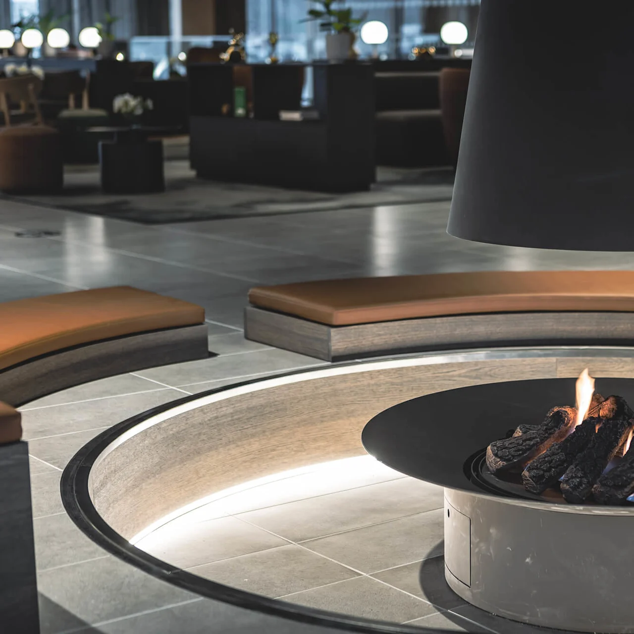 A circular fire pit emits flames in a modern indoor lounge. Surrounding it are curved seating areas with brown cushions, and background features soft lighting and contemporary furniture.