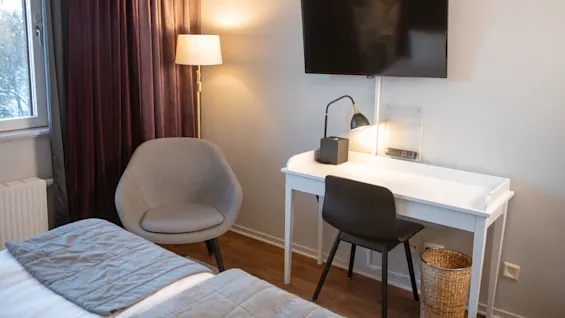 A wall-mounted TV is above a desk with a lamp and chair in a cozy room, featuring a gray armchair, floor lamp, and window with purple curtains.