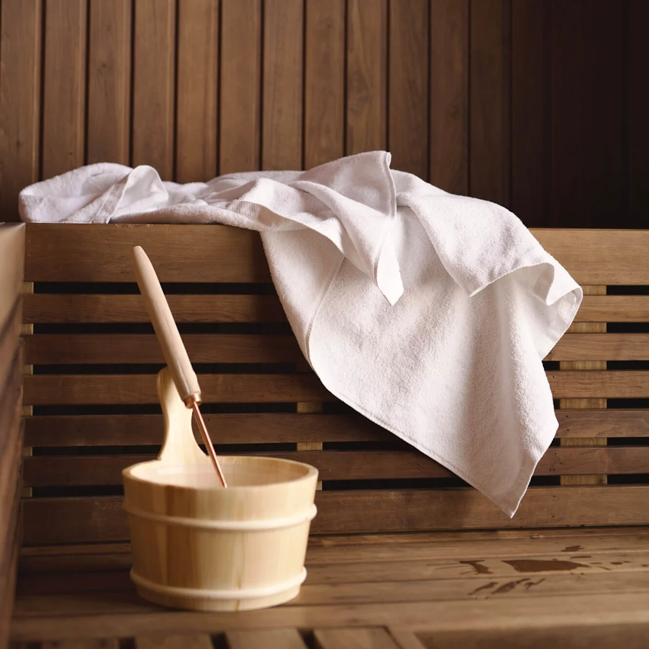 A wooden sauna bucket with a ladle beside a white towel, on wooden benches.