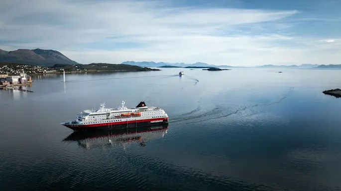 Cruise ship labeled "Hurtigruten" travels through calm water, leaving a wake. Mountains and a small coastal town are visible in the background beneath a partly cloudy sky.