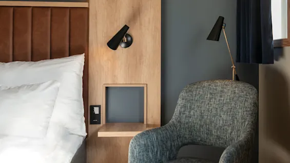 Säng med vita kuddar står mot en brun vadderad sänggavel. En träpanel med en svart vägglampa är intill. En texturerad stol står nära, under ett fönster med en gardin.