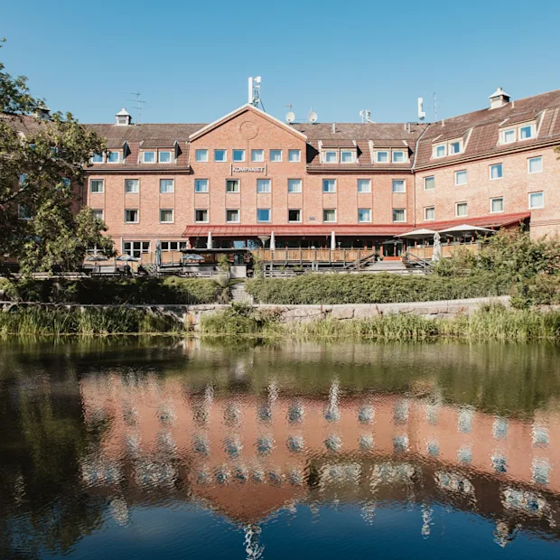 En stor tegelbyggnad med en skylt där det står KOMPANIET speglas i ett lugnt vattendrag med träd och grönska.