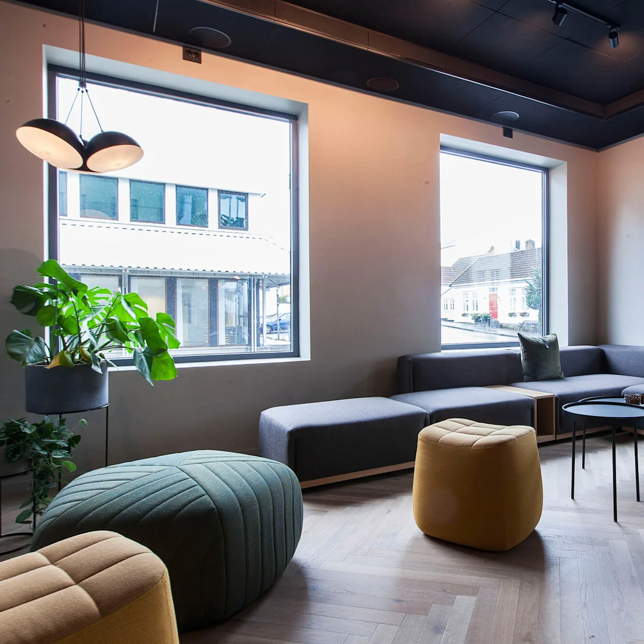 En grå modulsofa med grønne og gule puffer står i en moderne stue, omgitt av store vinduer og planter på parkettgulvet.