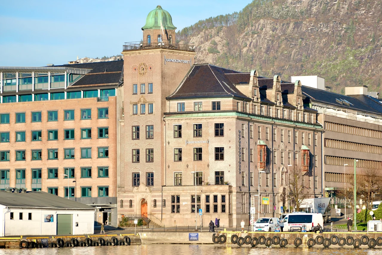 A charming waterfront scene featuring a historic building with a green dome and the text HAVNEKONTORET, against a mountain backdrop.
