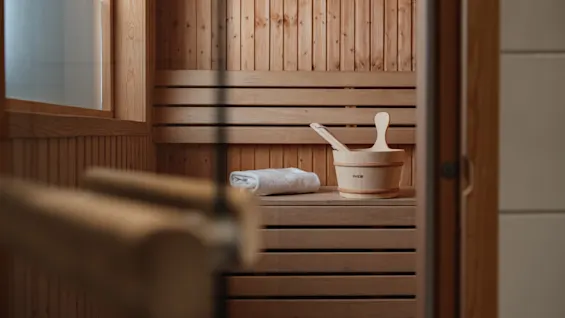Wooden sauna bench holds a rolled towel and a wooden bucket with a ladle, surrounded by paneled sauna walls.