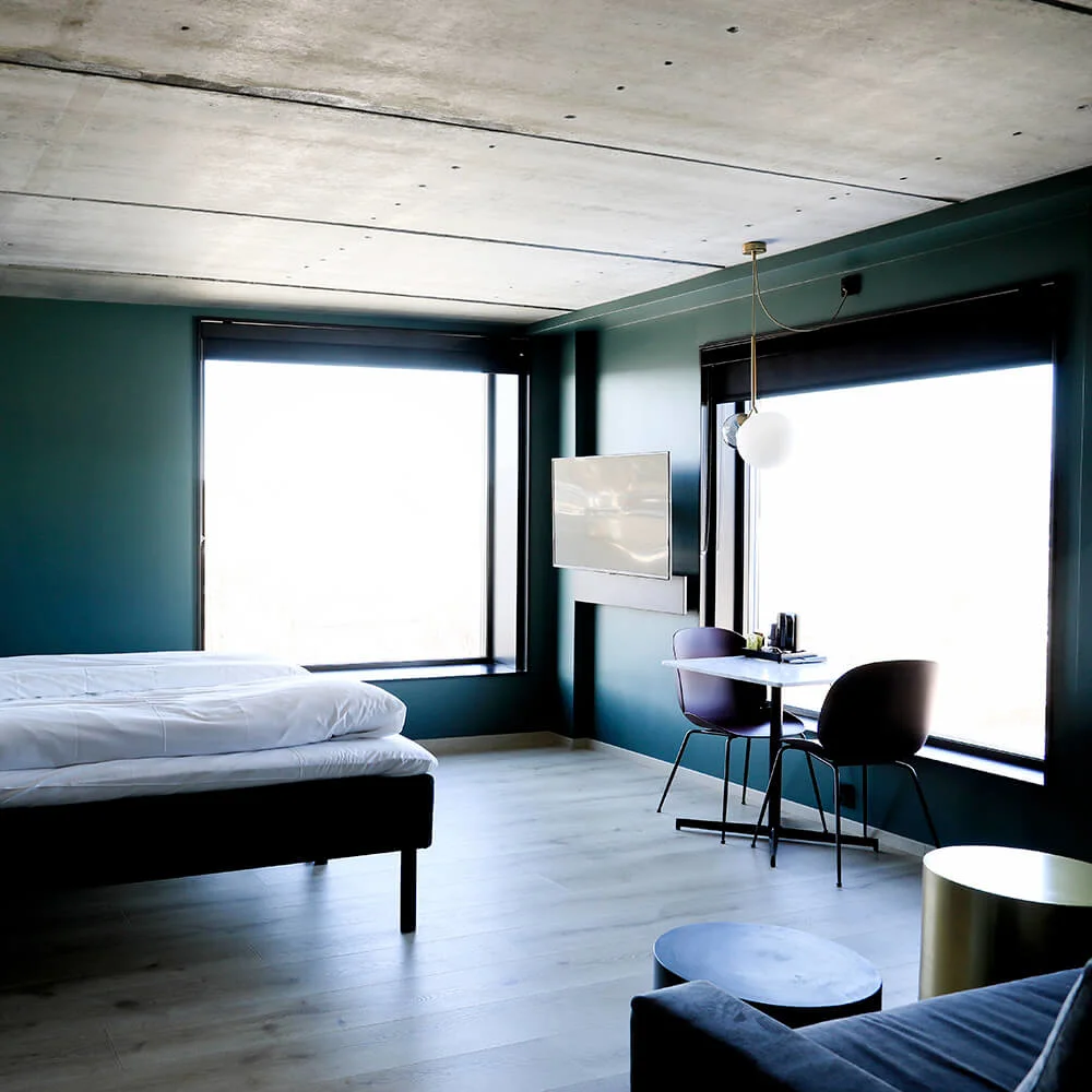 Et moderne hotelværelse med en seng placeret nær store vinduer. Et lille bord med stole står ved vinduet, og et fladskærms-TV er monteret på væggen.