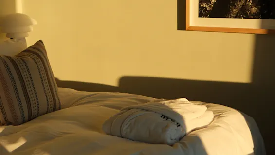 Varmt sollys oplyser en pænt redt seng med en foldet badekåbe og en dekorativ pude.