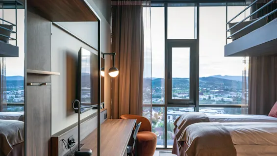Et hotellrom med et skrivebord i tre og lampe mot et stort vindu som viser byutsikt og fjell. To enkeltsenger med brune tepper står til høyre under myk belysning.