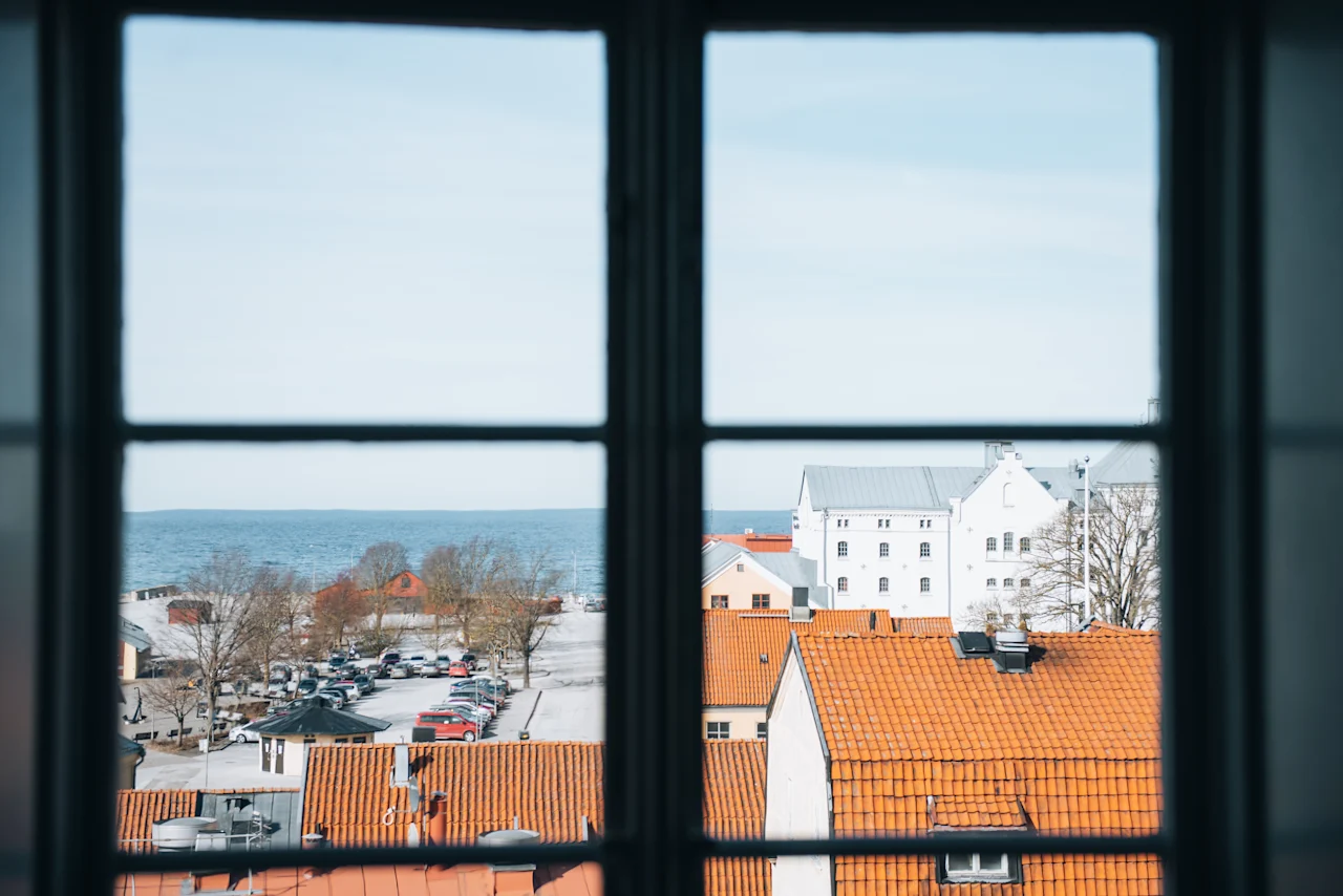 Udsigt gennem et vindue; orange-taget bygninger og parkerede biler i forgrunden. Træer og en stor vandmasse i det fjerne under en klar himmel.