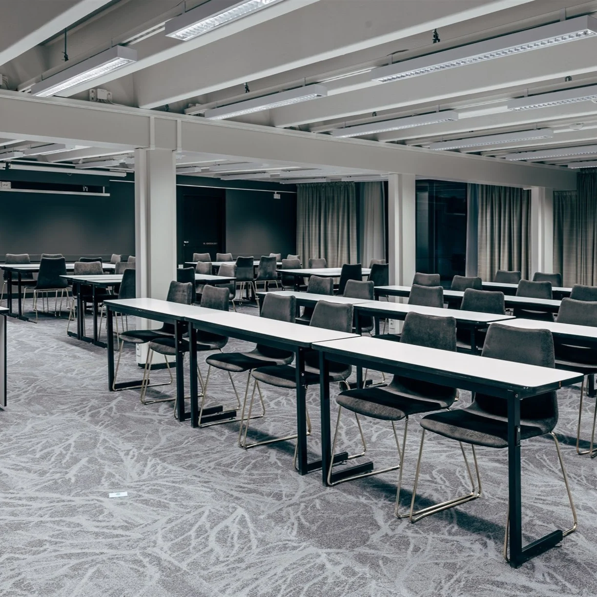 Long tables with black chairs are neatly arranged in a spacious conference room with grey carpeting. Large windows with curtains are on the side walls. Lighting is from ceiling fixtures.