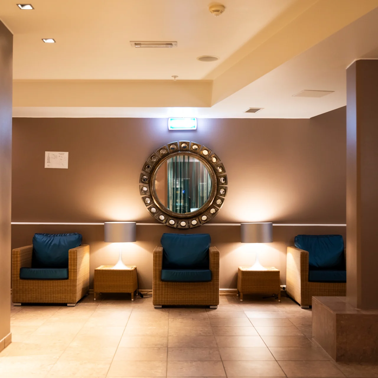 A round decorative mirror hangs on a brown wall, reflecting light above three blue-cushioned chairs placed evenly beneath it in a warmly lit lobby with tiled floors and columns. 