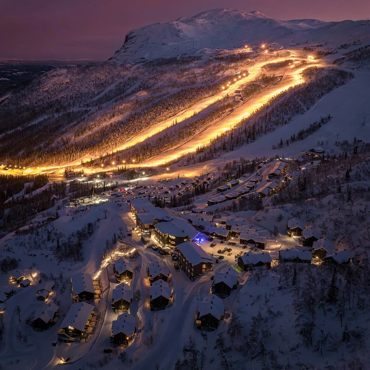 A brightly lit ski slope descends a snow-covered mountain at dusk, surrounded by a village with illuminated buildings nestled within a forested landscape.