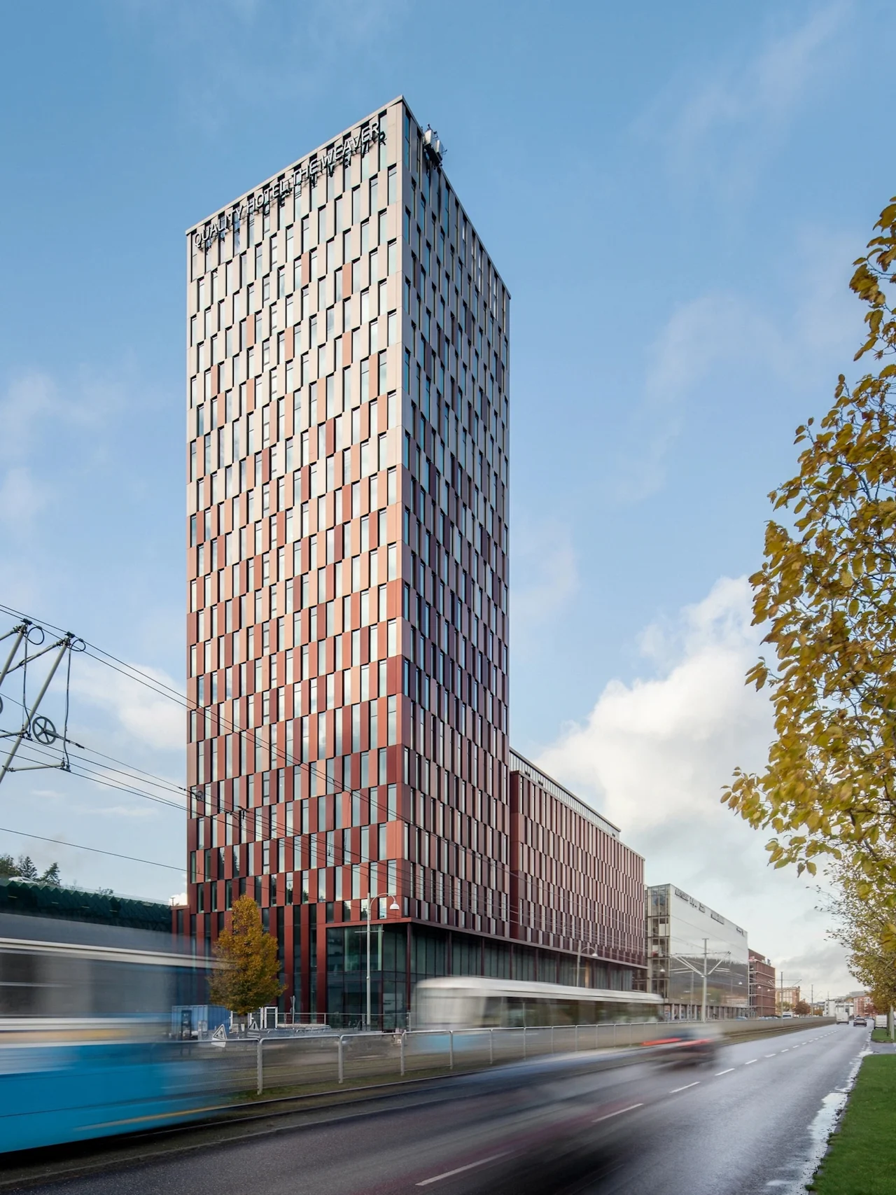 En modern hotellbyggnad med mönstrad fasad vid en livlig gata med suddiga spårvagnar och bilar, under blå himmel. Text: HOTEL THE WEAVER.