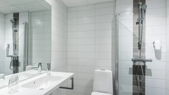 Sink beneath mirror with two glasses, adjacent to toilet; a shower is enclosed by glass doors, all in a white-tiled bathroom setting.