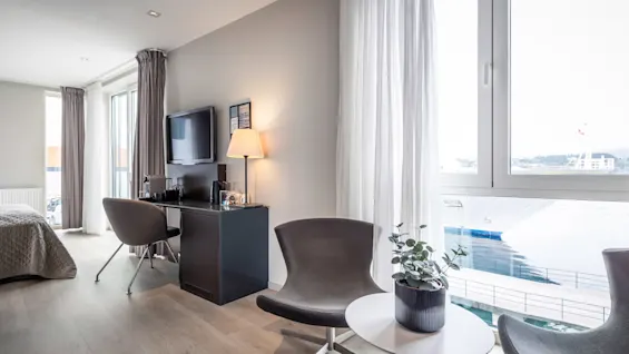 Ett modernt hotellrum har två stolar och ett litet bord med en växt vid ett fönster. En TV och lampa står på ett skrivbord, med en säng i bakgrunden.