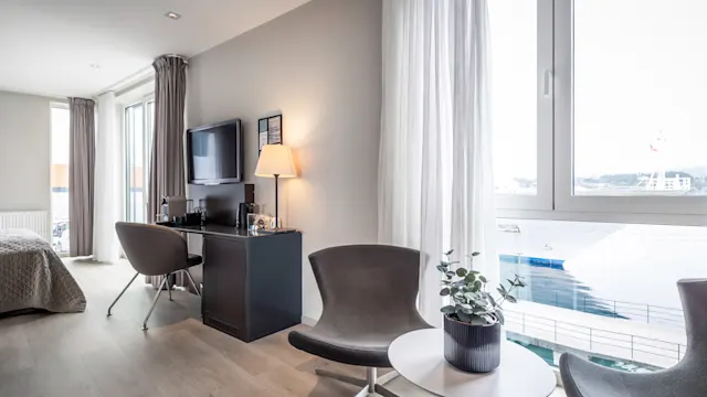 A modern hotel room has two chairs and a small table with a plant by a window. A TV and lamp are on a desk, with a bed in the background.