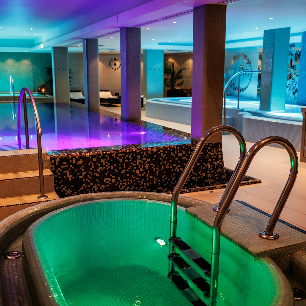 A jacuzzi with green lighting, next to a larger pool with purple illumination, in a modern spa environment featuring columns and ambient ceiling lights.