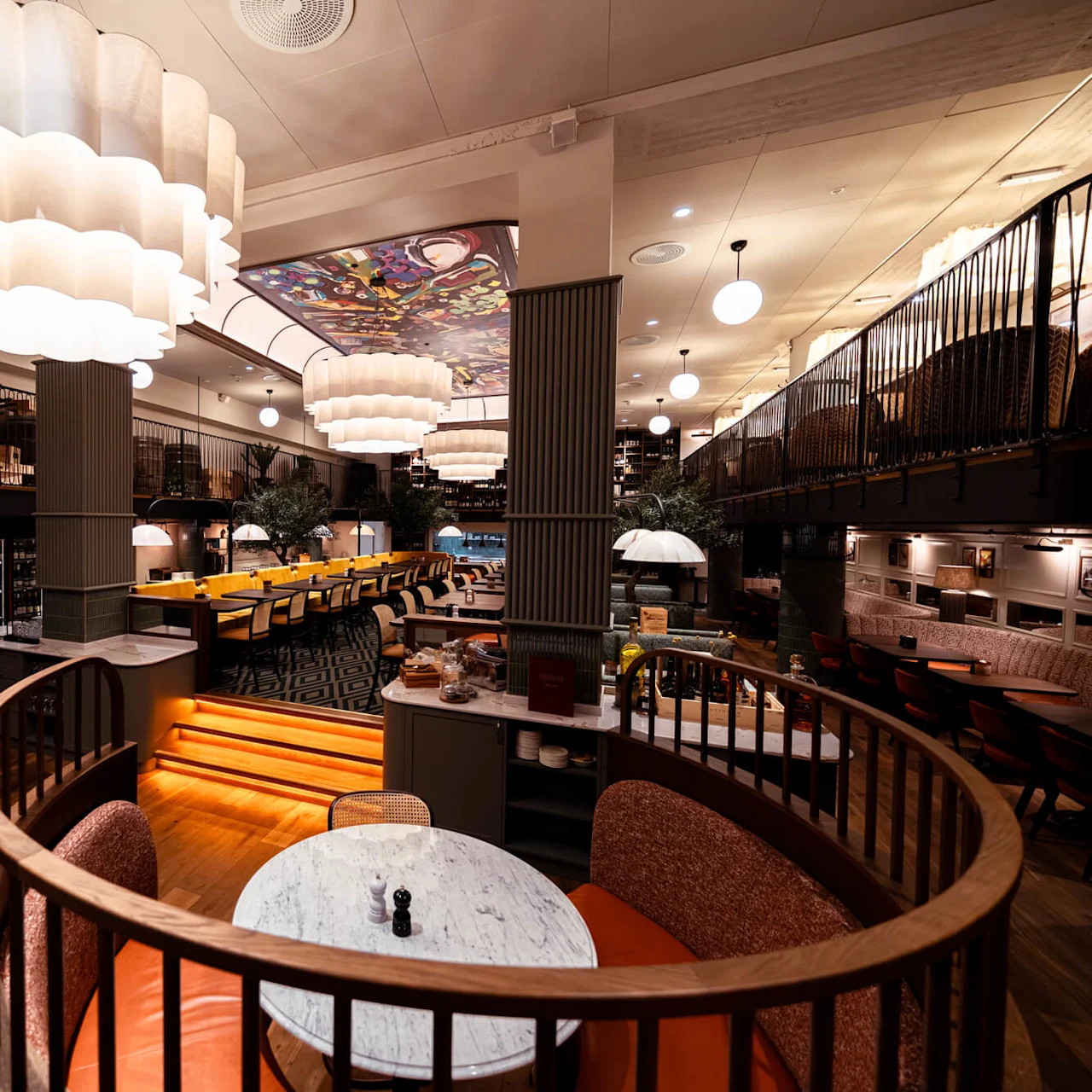 A restaurant interior featuring round booths and a central staircase. Overhead, large, wavy pendant lights illuminate the space. Surrounding walls display framed artwork, contributing to a cozy ambiance.