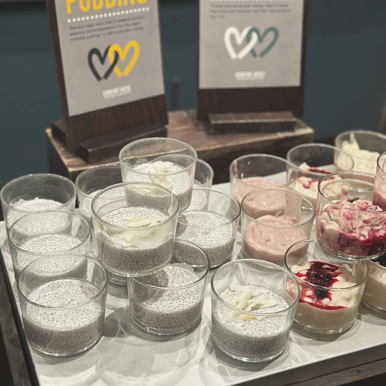 Glasses filled with chia pudding and over-night oats are arranged on a table. Signs above them read "CHIA PUDDING" and "OVER-NIGHT OATS" with descriptive text and logos.