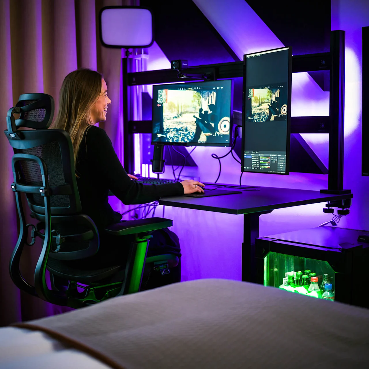 A person sits in a black ergonomic chair, gaming on dual monitors, illuminated by purple lights in a modern room with a mini-fridge containing drinks.