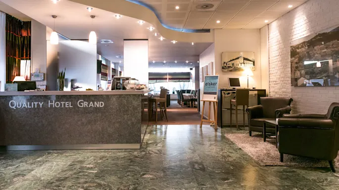 Reception desk with "QUALITY HOTEL GRAND" inscription; lounge area with armchairs and table. Illuminated interior; marble floor; poster with "elsker" text on easel, lamps and wall art visible.