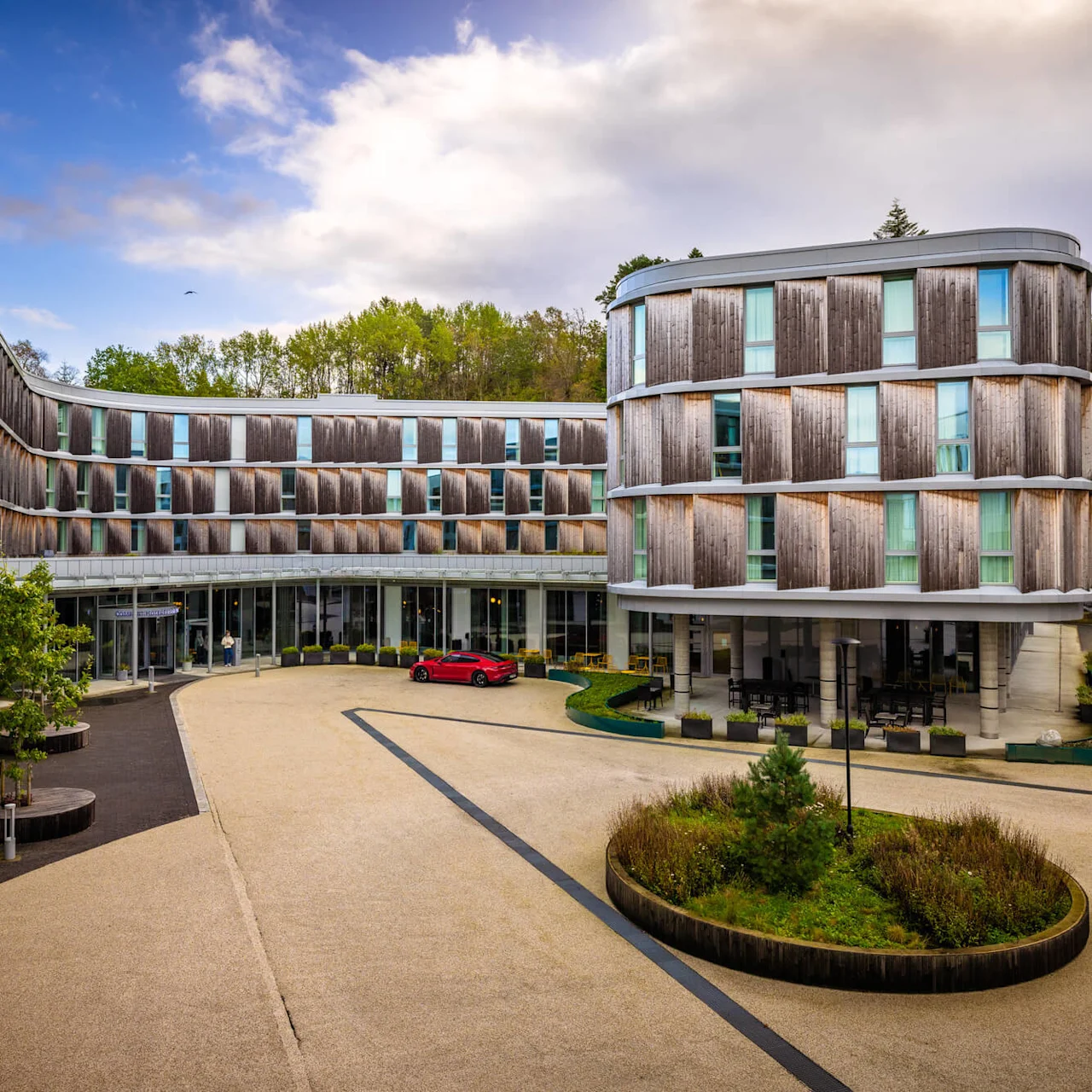 A modern hotel building with wooden panels stands in a courtyard with a central green island and a red car parked. Forest trees are visible in the background.