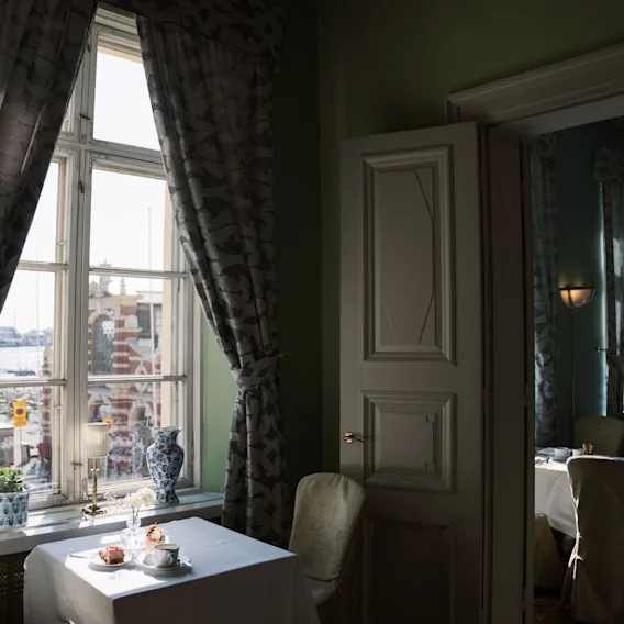 Table set with cups and pastries sits by a window with flowing curtains. Soft light fills the room, revealing an elegant interior and outdoor view of buildings and water.