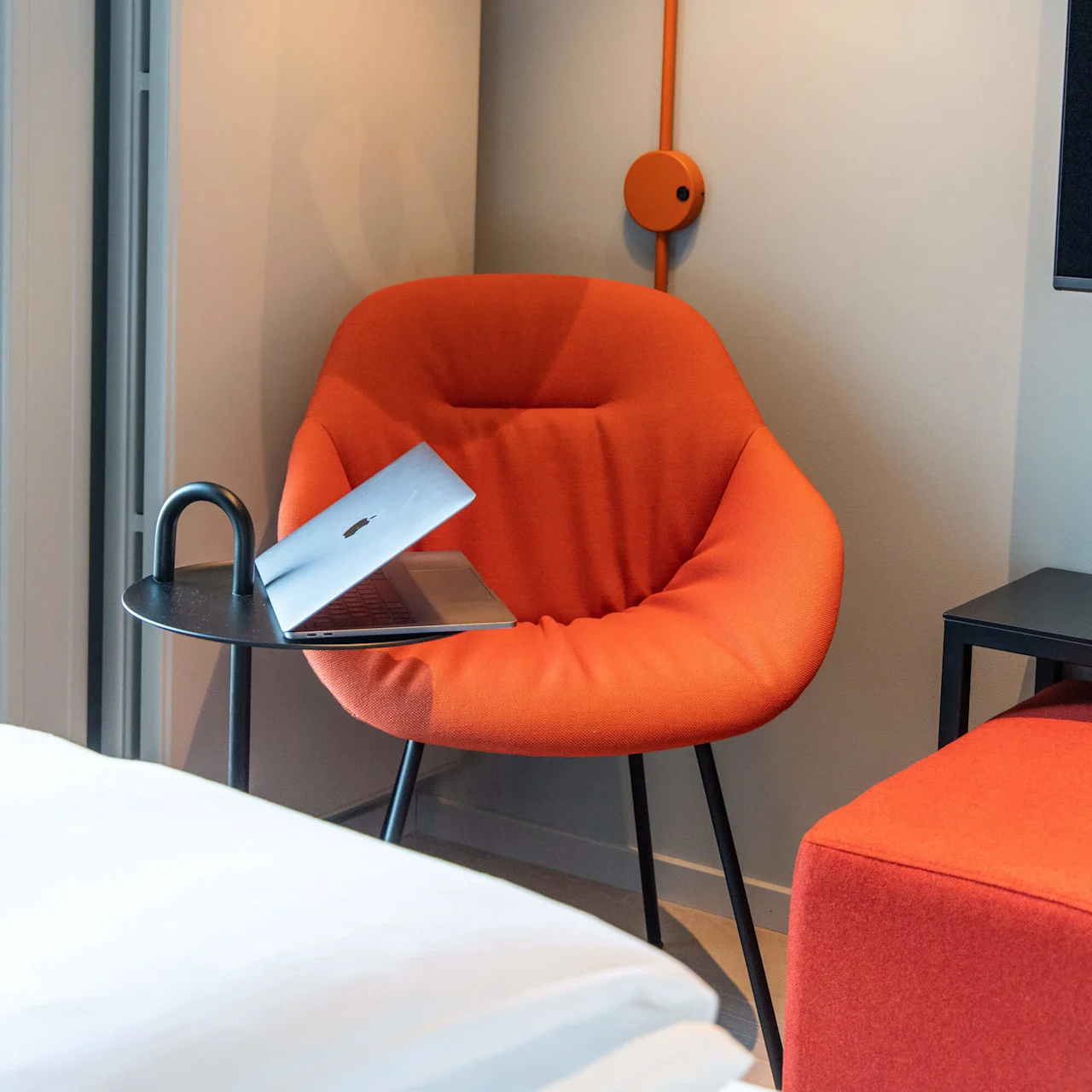 An orange chair holds a partially closed laptop, beside a small black side table. A matching wall lamp hangs above, in a minimalist room.
