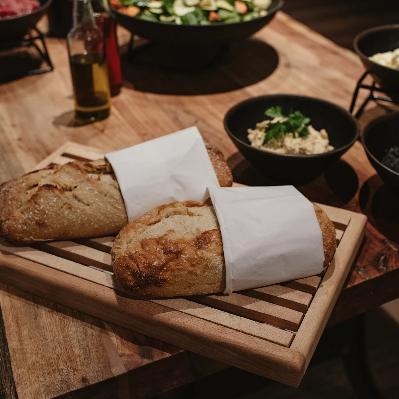 Two loaves of bread wrapped in paper rest on a wooden cutting board. Surrounding them are bowls of various appetizing dishes on a rustic wooden table.