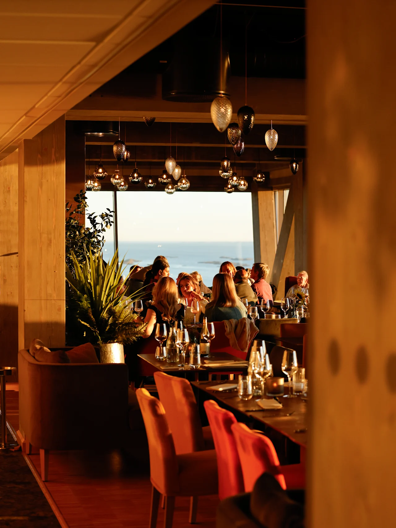 En gruppe mennesker spiser ved elegant dækkede borde i en varmt oplyst restaurant med store vinduer, der giver udsigt til havet, under et loft med hængende lamper.