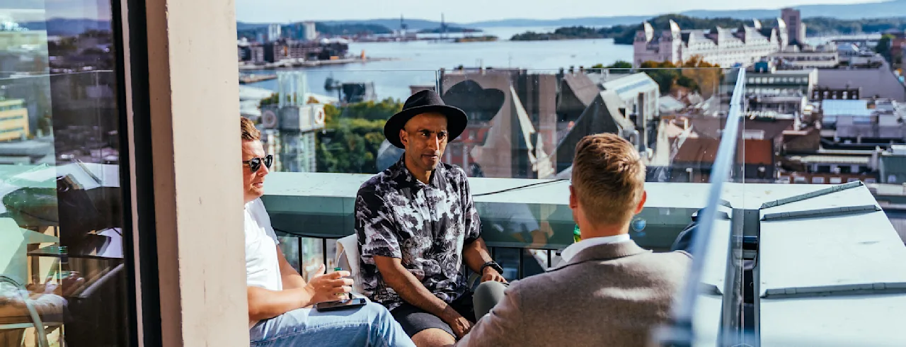 Kolme ihmistä istuu keskustelemassa kattoterassilla, joka avautuu näkymään kaupunkiin ja joen ylle aurinkoisena päivänä.