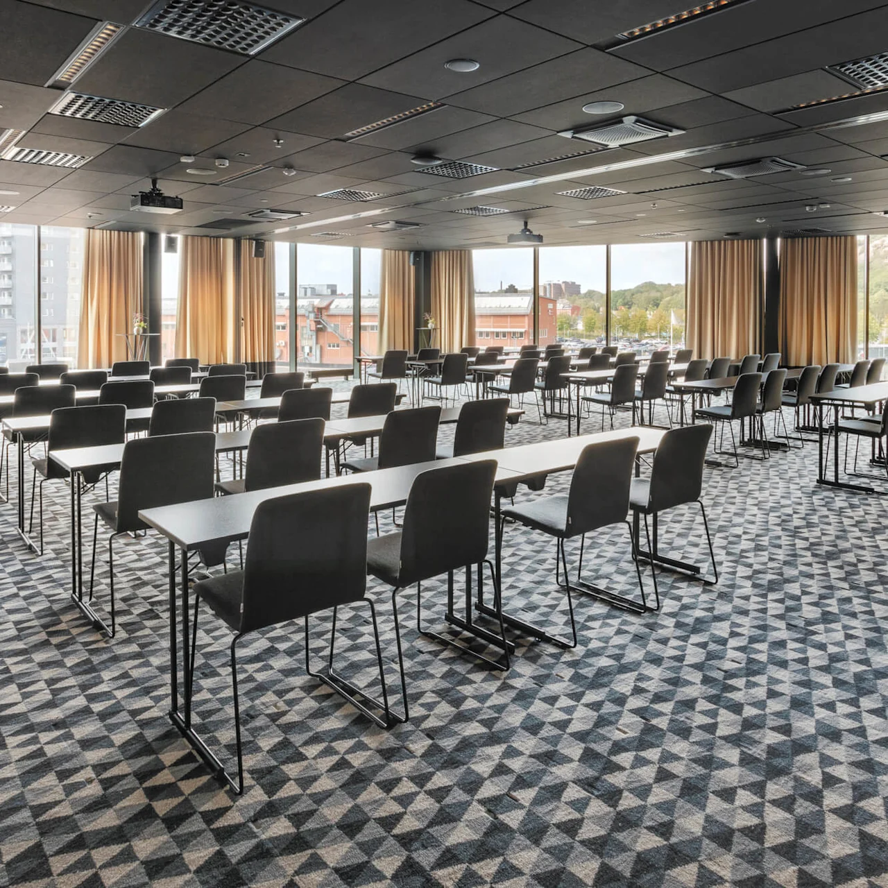 Ett rymligt konferensrum har rader av tomma svarta stolar och bord. Golv-till-tak-fönster med gardiner erbjuder naturligt ljus och stadsutsikter. Geometrisk matta täcker golvet.
