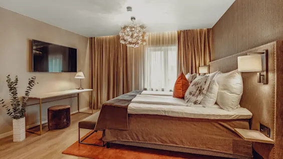 A neatly made bed in a well-lit, cozy hotel room with modern décor, a flat-screen TV, and elegant curtains.