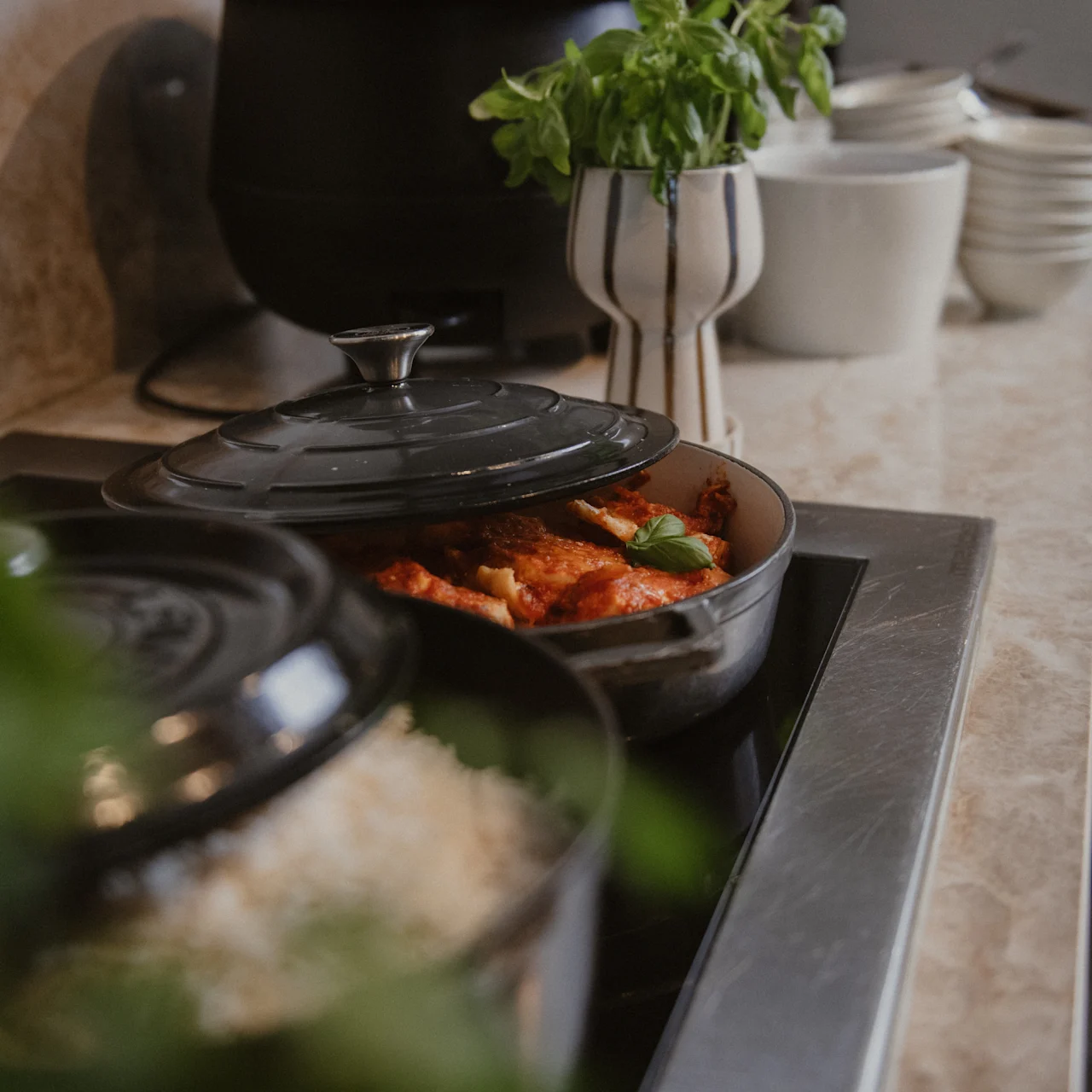 Pot on stove, simmering food under a lid, with rice in another pot nearby. Kitchen counter displays stacked plates and bowls beside a vase of fresh herbs.