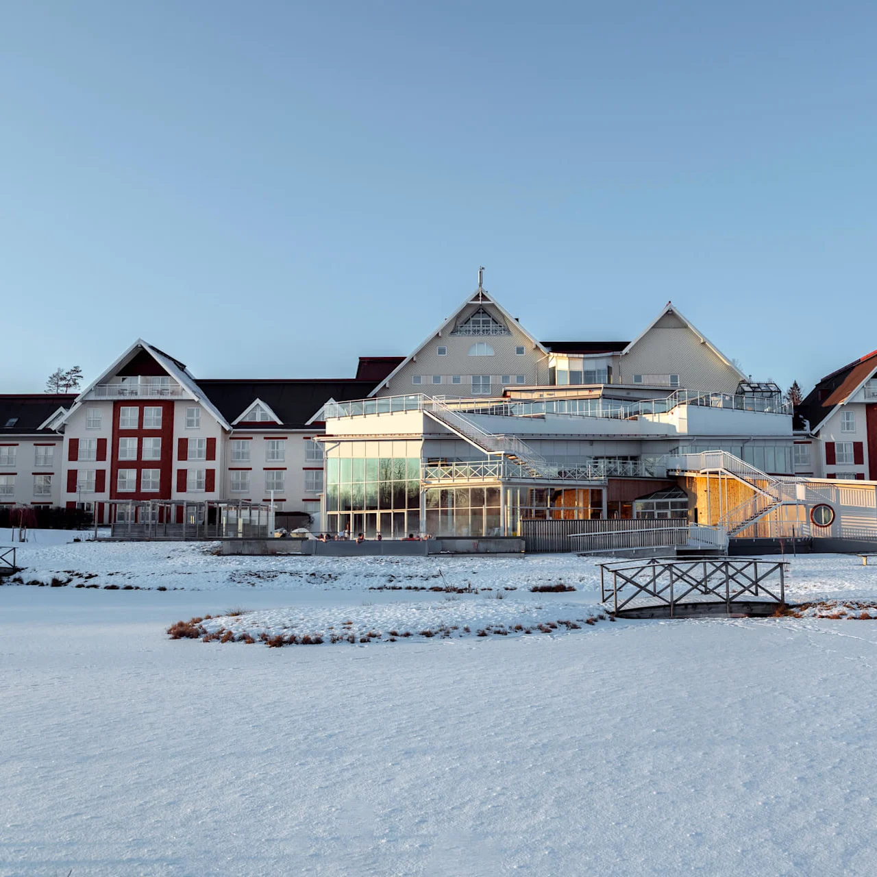 En stor, hvit og rød hotellbygning står under en blå himmel, omgitt av snødekt bakke og en frossen innsjø, med trær i bakgrunnen.