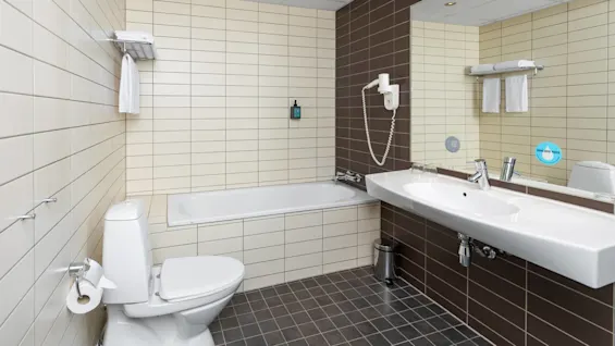 Toilet, bathtub, and sink are arranged in a tiled bathroom, with towels on a rack and a wall-mounted hairdryer. A sign reads "DRINK MORE WATER."