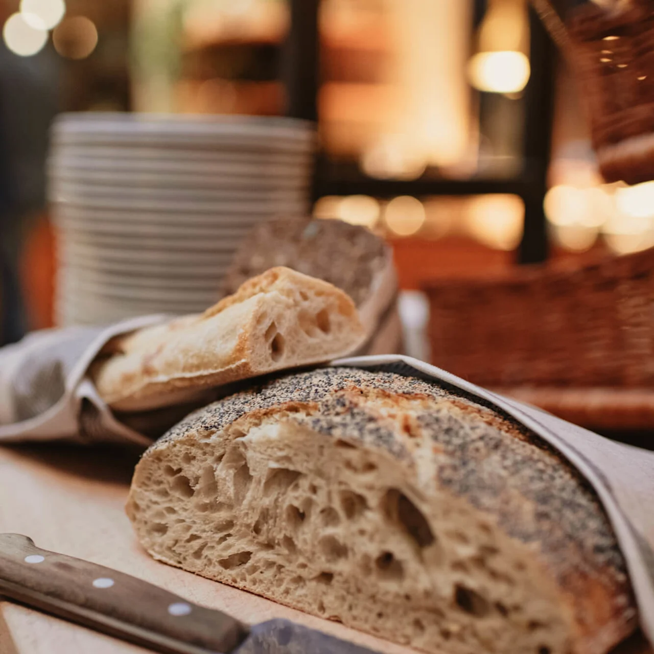 Bread slices rest on a wooden board beside a knife, surrounded by baskets and stacked plates in a warm, softly lit environment.