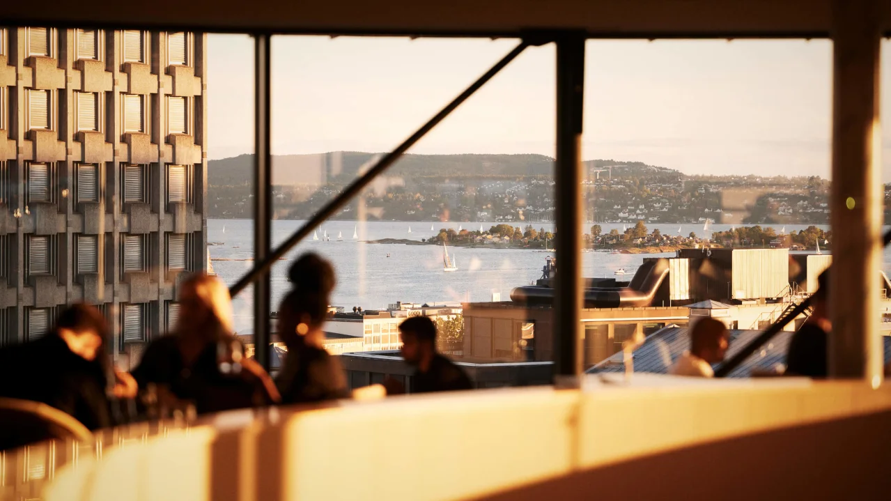 People sit in an indoor space with large windows overlooking a sunlit cityscape and water with boats.