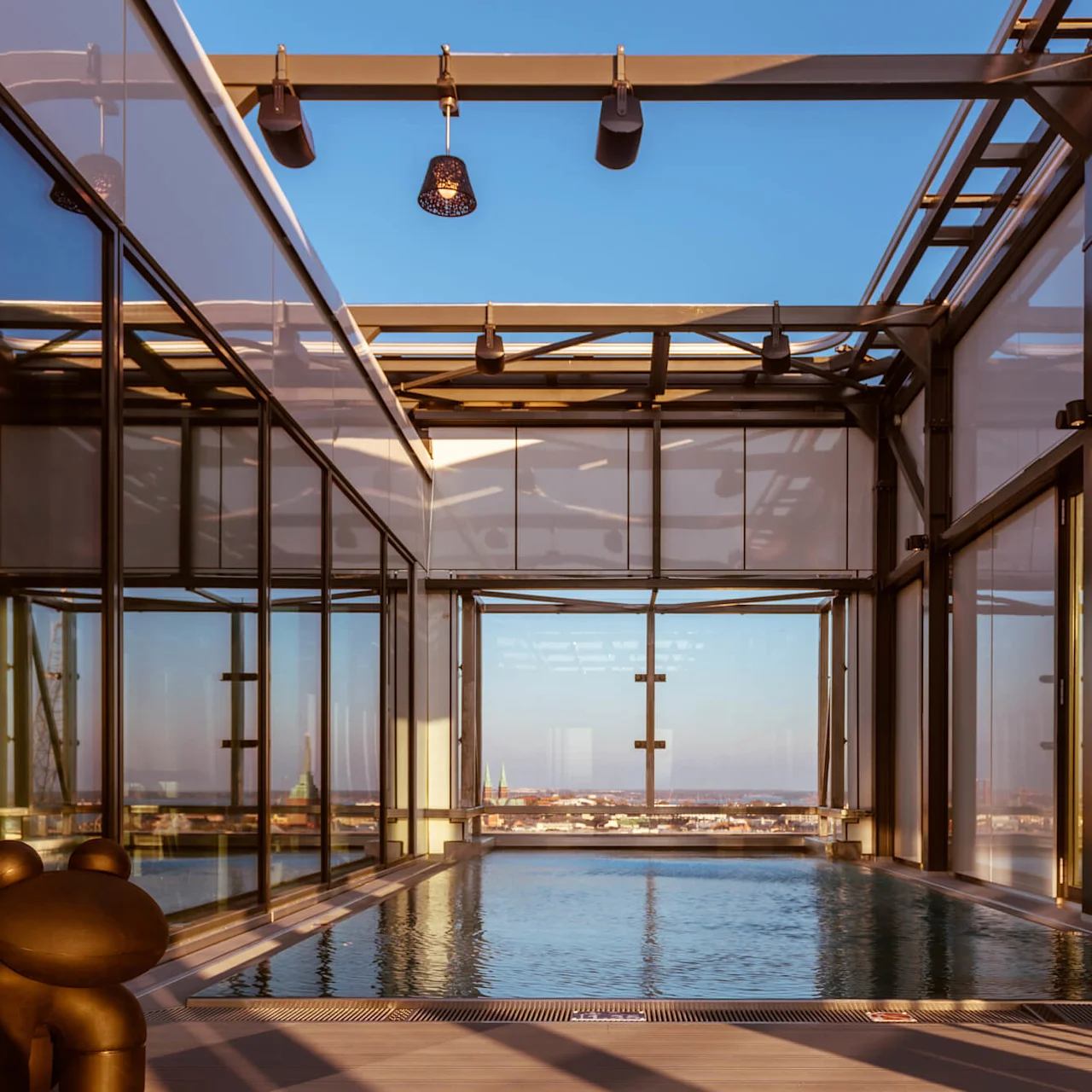 En moderne indendørs pool strækker sig under et glastag og tilbyder udsigt over en bysilhouet i det fjerne. Geometriske strukturer og kunstnerisk belysning fremhæver den luksuriøse atmosfære.