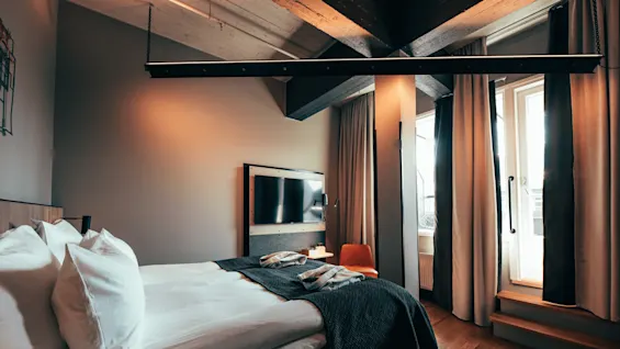 En pent oppredd seng med sammenbrettede håndklær, plassert i et moderne rom med en veggmontert TV. Et vindu med forheng slipper inn mykt naturlig lys.