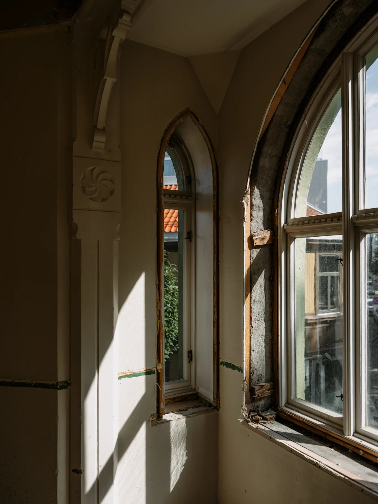 Sollys strømmer ind gennem to buede vinduer i et rum under renovering og kaster dramatiske skygger på væggene.