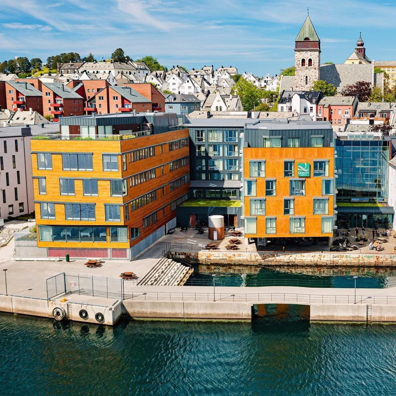 Ett komplex vid vattnet har moderna orangea och glasade byggnader vid en kanal. I bakgrunden ligger en pittoresk stad med ett framträdande kyrktorn under en blå himmel.